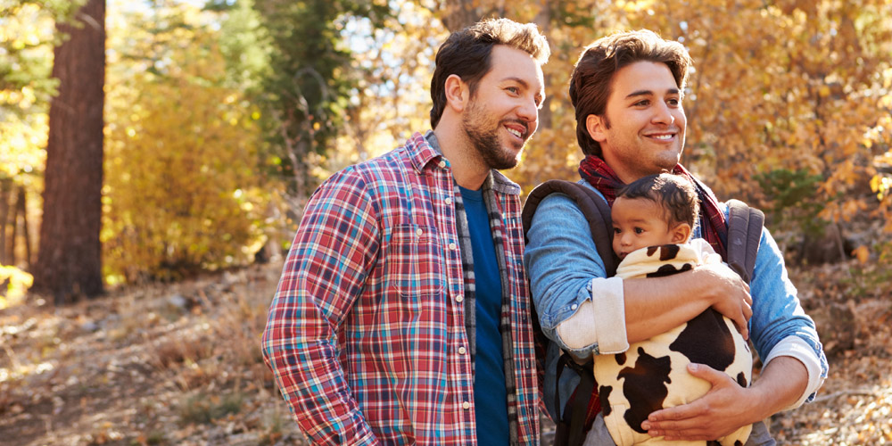 A same-sex male couple walking together among the fall foliage with one wearing a baby in a front carrier