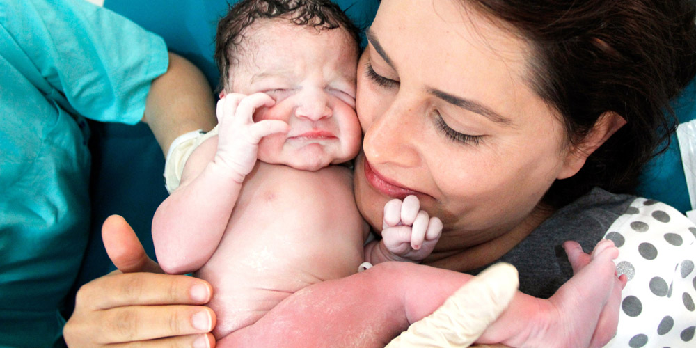 A woman who just gave birth reaching out to hold her newborn, who is in the hands of a nurse or doctor 
