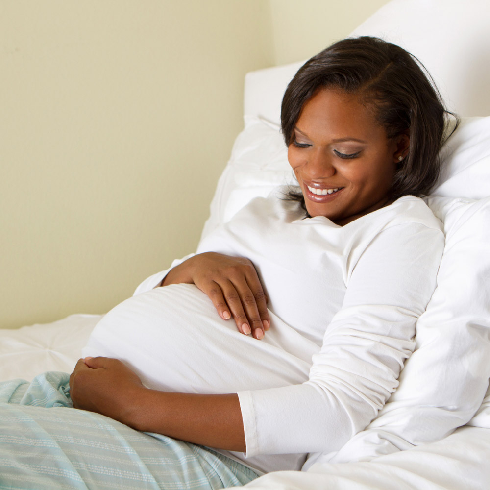 An African-American pregnant woman sitting, smiling, on a bed, holding her belly in her hands